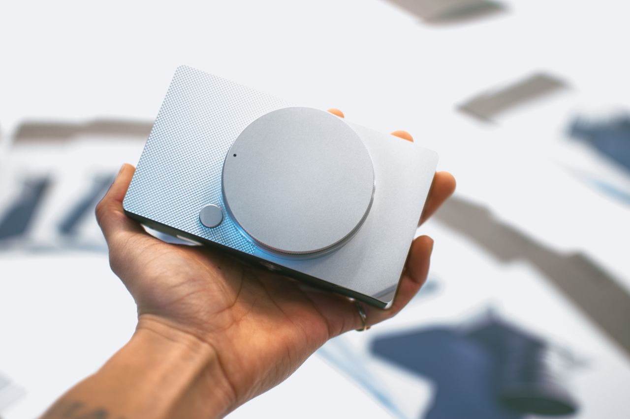 Home Close-up of a hand holding a sleek gray portable speaker against a blurred background.